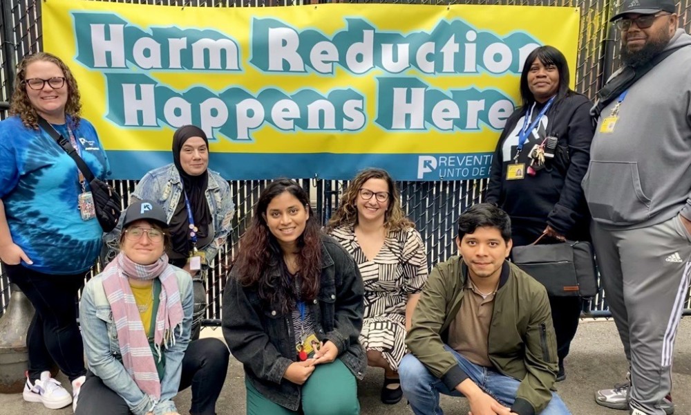 8 staff smile at the camera in front of a banner that reads, "Harm Reduction Happens Here."