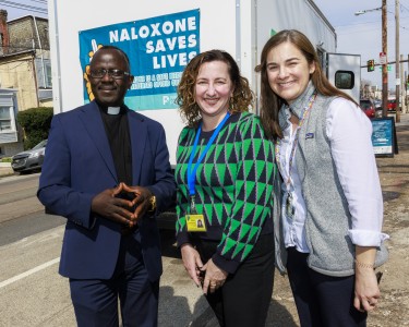 Reverend Father MacIvan Rogers, Director of Behavioral Health Denise Botcheos, and Dr. Judy Chertok stand in front of a bus with a banner reading "Naloxone Saves Lives"