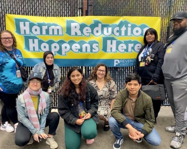 8 staff smile at the camera in front of a banner that reads, "Harm Reduction Happens Here."